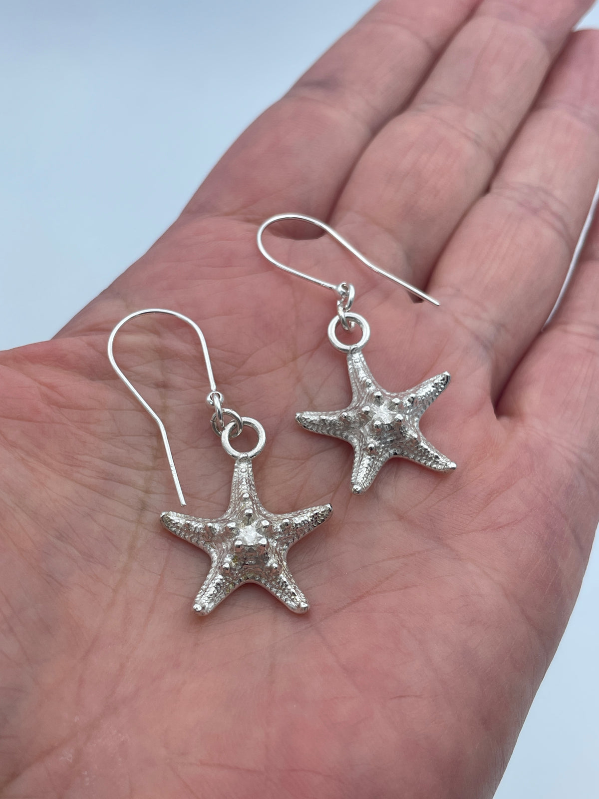 A pair of silver starfish drop earrings displayed on a person's palm against a light blue background.