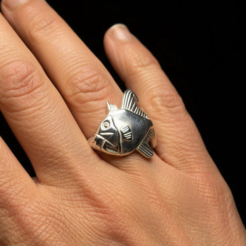 A silver ring with triggerfish design worn on a person's ring finger, shown against a plain light gray background.