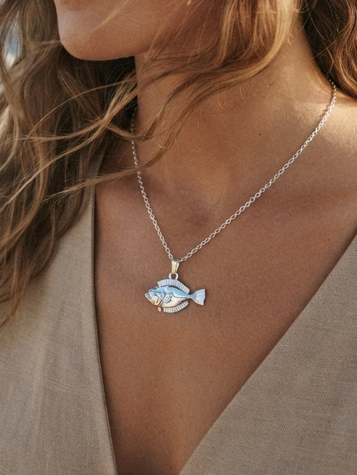 A woman wearing a silver fish pendant necklace, with the blue ocean and sky in the background.