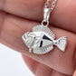 A silver fish pendant necklace held in a person's hand. The pendant is detailed with textured scales and a matching silver chain is visible at the top.