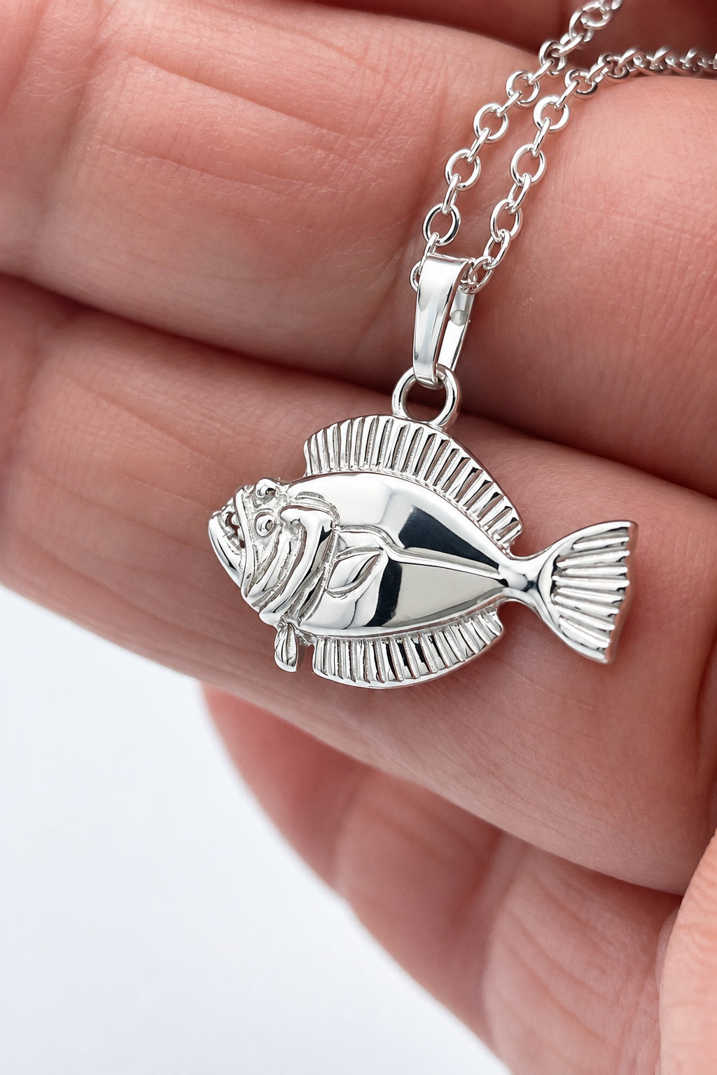 A silver fish pendant necklace held in a person's hand. The pendant is detailed with textured scales and a matching silver chain is visible at the top.