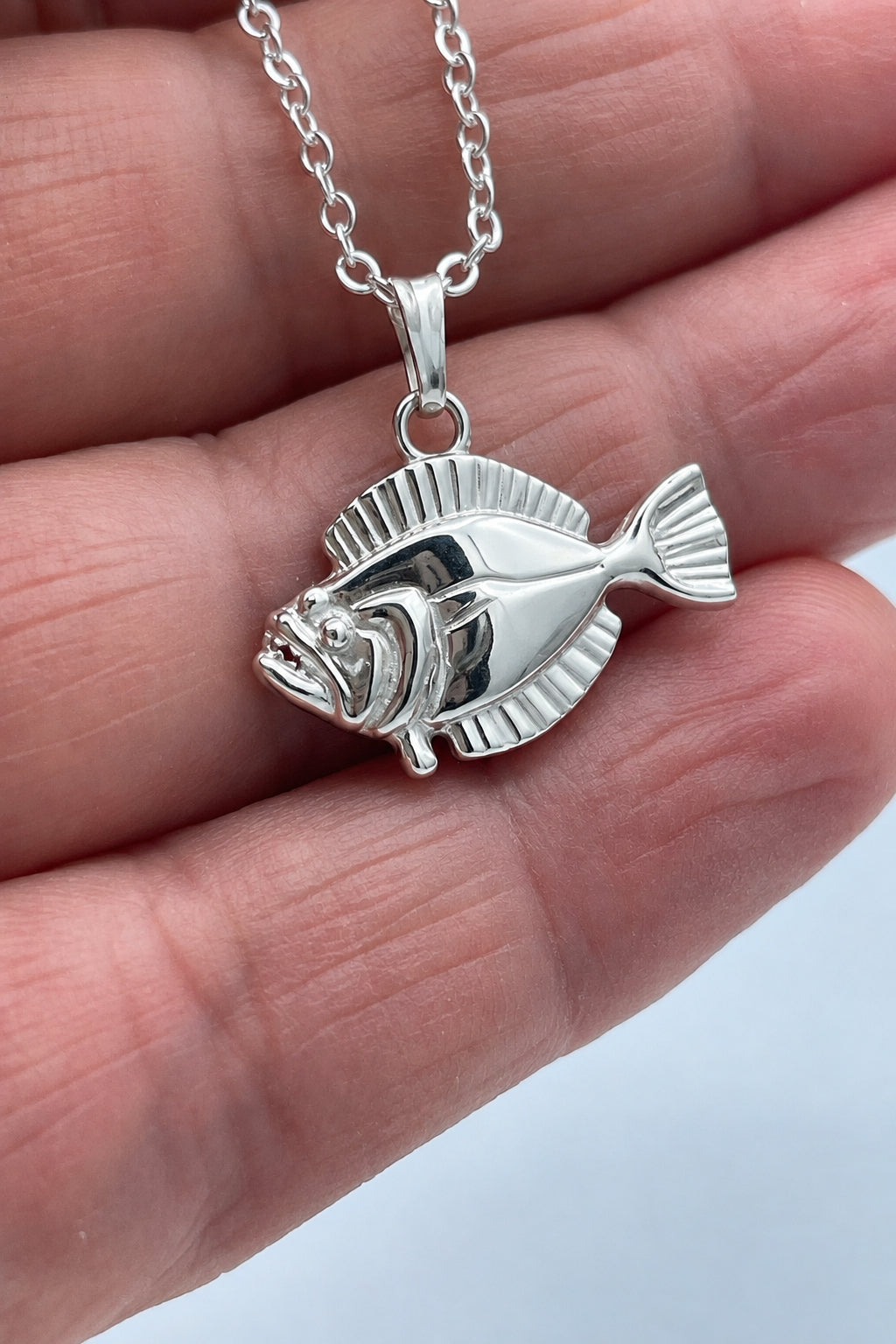 A silver fish pendant necklace held on a person's fingers against a light gray background.