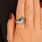 A close-up of a hand wearing a silver ring shaped like a freshwater trout, set against dark black fabric.