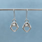 A pair of silver drop earrings shaped like shark teeth, hanging from a textured metal rod against a soft blue-gray background.