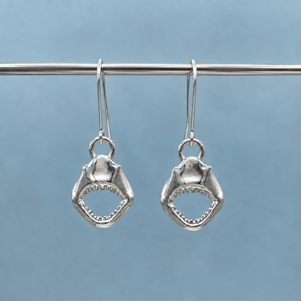 A pair of silver drop earrings shaped like shark teeth, hanging from a textured metal rod against a soft blue-gray background.