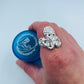 A hand wearing a large, textured silver ring with a coral or sea urchin-like pattern, holding a blue bottle labeled 'Atlantis Ocean Jewelry' against a light blue background.