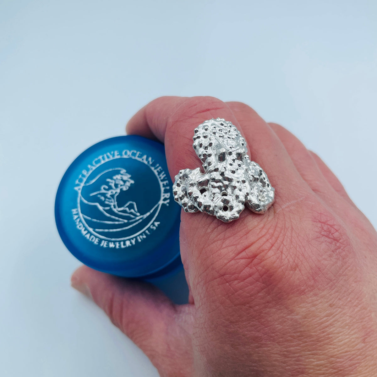 A hand wearing a large, textured silver ring with a coral or sea urchin-like pattern, holding a blue bottle labeled 'Atlantis Ocean Jewelry' against a light blue background.