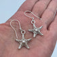A pair of silver starfish drop earrings displayed on a person's palm against a light blue background.