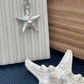A silver starfish pendant necklace on a matching chain, styled on a white ribbed surface next to a real white starfish and a wood-grain background.