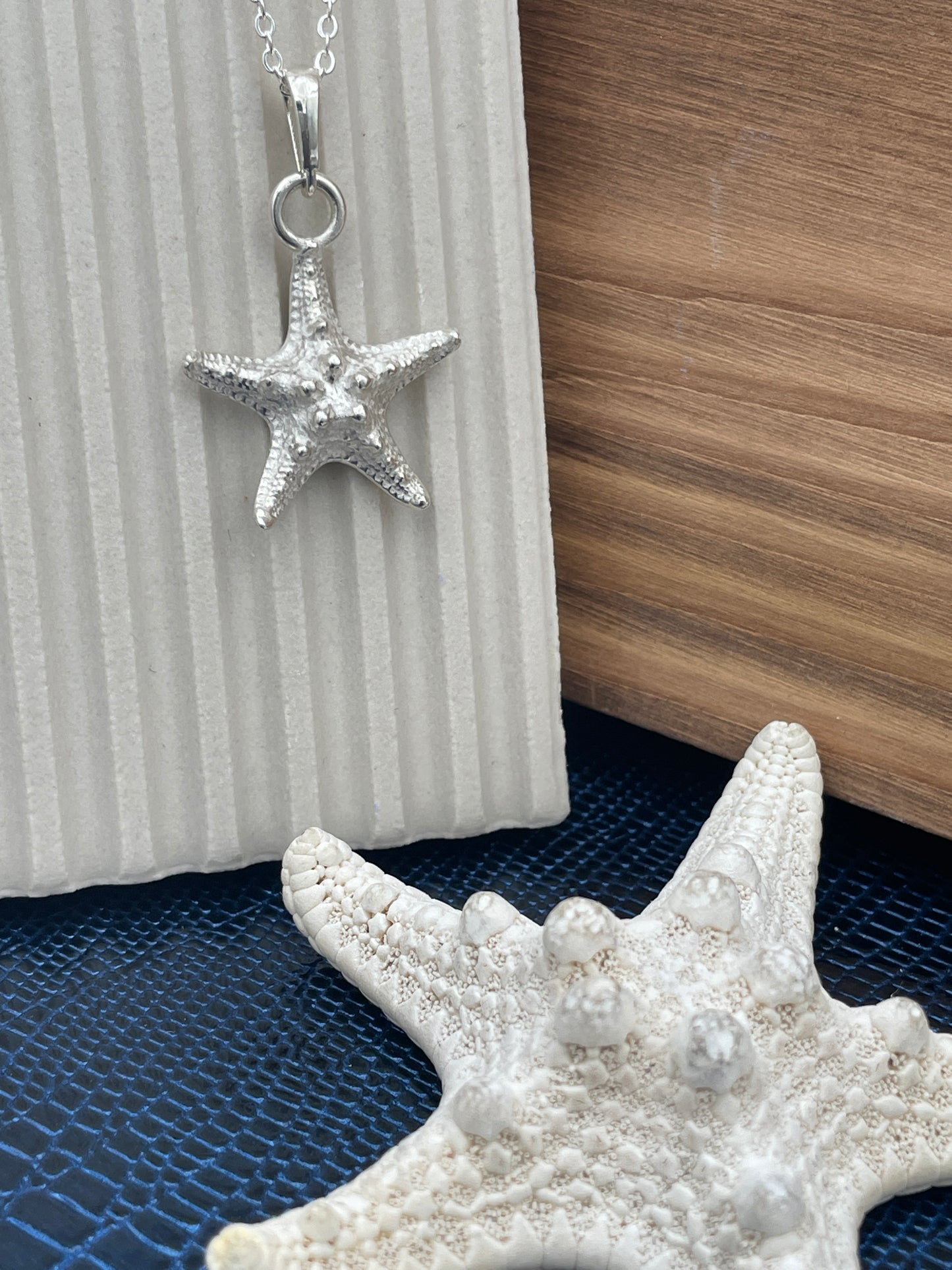 A silver starfish pendant necklace on a matching chain, styled on a white ribbed surface next to a real white starfish and a wood-grain background.